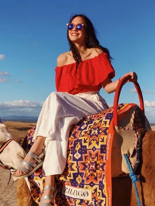 Duduk di atas unta, Pevita memesona dalam balutan outfit merah yang kasual. Atasan off-the-shoulder cropped top dipadunya dengan celana panjang putih, sandal silver, dan sunglasses. Foto: Instagram.