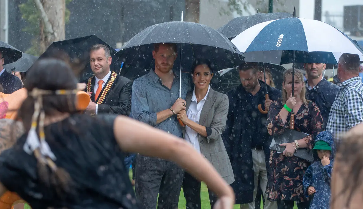 Pangeran Harry dan Meghan Markle berlindung di bawah payung menyaksikan tarian aborigin saat piknik di Victoria Park, Dubbo, Australia, Rabu (17/10). Keduanya diagendakan tur ke Australia dan Pasifik Selatan selama 16 hari. (Ian Vogler/Pool via AP)