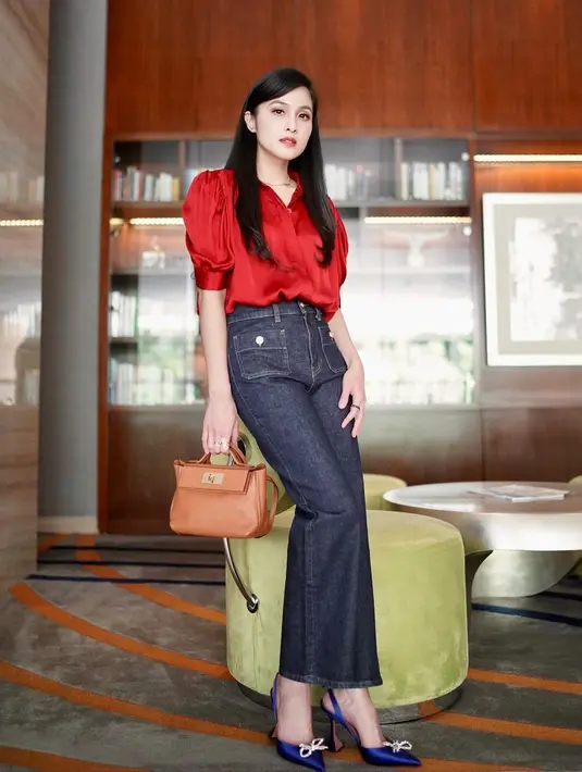 Sandra Dewi tampil menawan dalam balutan top blouse merah dipadunya dengan denim pants, menenteng handbag cokelat, dan high heels biru navy. [Foto: Instagram/sandradewi88]