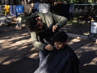 Mohammed al-Hamo (kiri) memotong rambut saudara laki-lakinya, Sobhi di depan tenda mereka di kamp darurat pengungsi korban gempa Turki dan Suriah di kota Antakya pada 19 Februari 2023. Mohammed al-Hamo tak pernah membayangkan akan menggunakan keterampilannya mencukur rambut akan digunakan di tenda pengungsi korban gempa Turki dan Suriah. (Sameer Al-DOUMY / AFP)