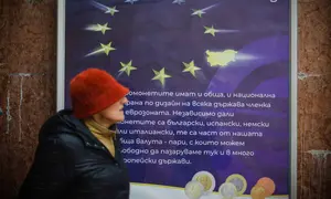 Mengadopsi euro dinilai sebagai fase akhir integrasi Bulgaria ke arus utama Eropa, setelah bergabung dengan NATO, Uni Eropa, kawasan Schengen, dan kini zona mata uang tunggal. Tampak dalam foto, seorang wanita berjalan melewati poster yang menampilkan Bulgaria sebagai anggota Zona Euro ke-21, di Sofia, Kamis 1 Januari 2026, saat negara tersebut bergabung dengan serikat mata uang euro. (AP Photo/Valentina Petrova)
