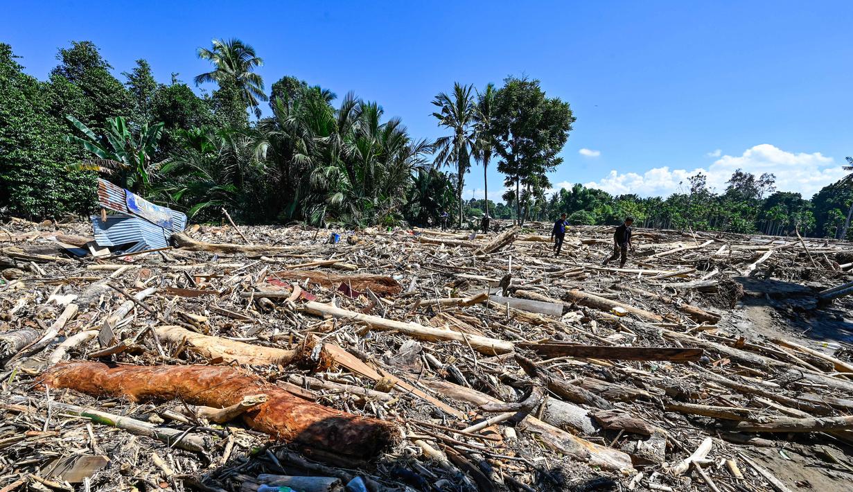 Pemerintah Provinsi Aceh menyebutkan, bencana kali ini menjadi yang terparah setelah gempa dan tsunami 2004. Tampak dalam foto, orang-orang berjalan melintasi lumpur dan puing-puing di daerah terdampak banjir di Meureudu, Kabupaten Pidie Jaya, Provinsi Aceh, pada 30 November 2025. (CHAIDEER MAHYUDDIN/AFP)