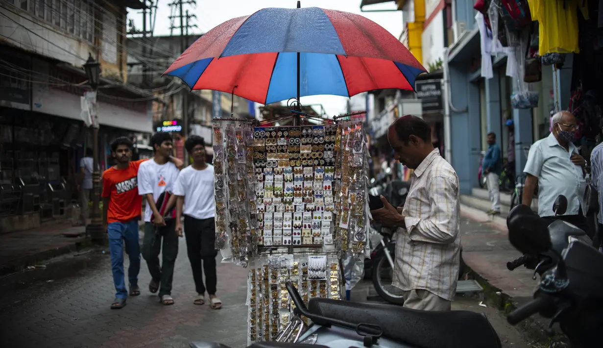 Seorang pedagang kaki lima menggunakan payung untuk melindungi anting-antingnya dari hujan di Kochi, negara bagian Kerala, India, Rabu (6/7/2022). Negara bagian selatan itu telah mewajibkan penggunaan masker di tempat-tempat umum menyusul meningkatnya kasus COVID-19. (AP Photo/ RS Iyer)