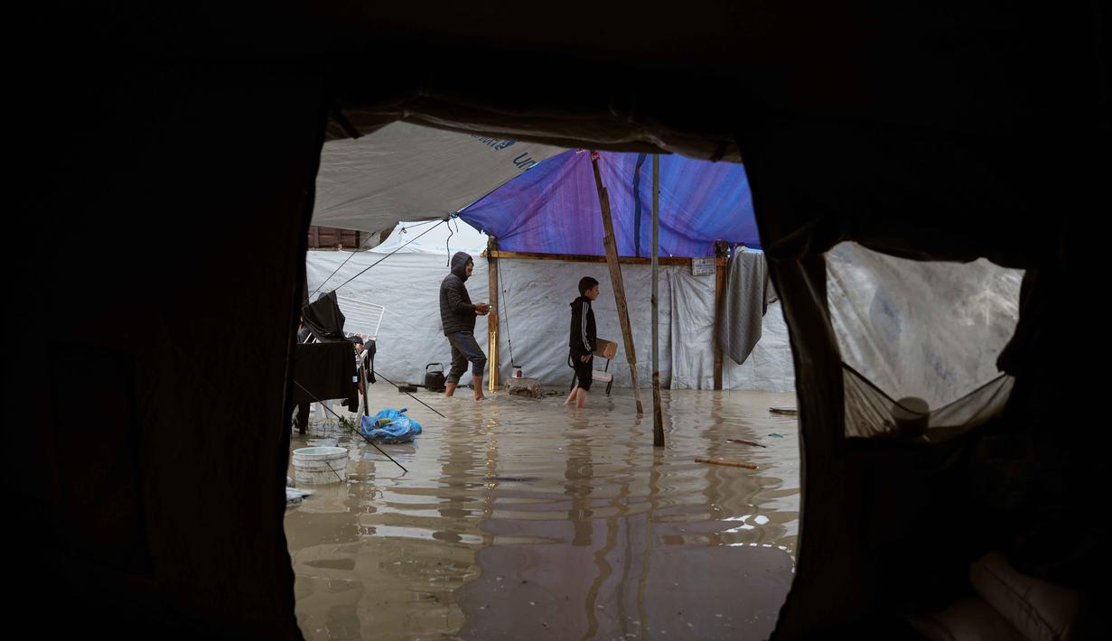 Para pengungsi Palestina berjalan melewati kamp tenda yang basah kuyup akibat hujan lebat di Deir al-Balah, Jalur Gaza tengah, Kamis, 26 Maret 2026. Kondisi kamp pengungsian di Gaza sangat memprihatinkan akibat banjir yang terus berulang selama musim dingin. (AP Photo/Abdel Kareem Hana)