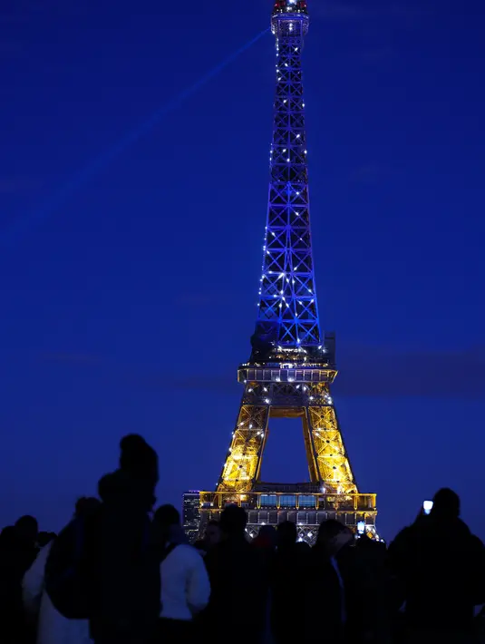 Warga melihat Menara Eiffel menyala dengan warna bendera Ukraina di Paris (25/2/2022). Pasukan Rusia menyerang ibu kota Ukraina pada Jumat (25/2) dengan tembakan dan ledakan dalam invasi ke negara demokratis yang memicu kekhawatiran perang yang lebih luas di Eropa. (AFP/ Thomas Coex)