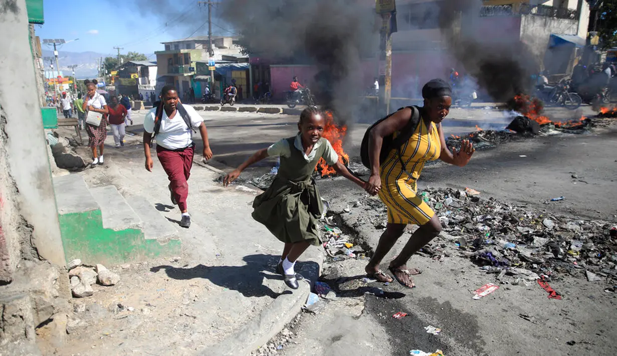 Seorang wanita dengan putrinya berjalan melewati barikade yang didirikan oleh anggota polisi saat mereka memprotes tata kelola polisi yang buruk di Port-au-Prince, Haiti, 26 Januari 2023. Banyak bisnis dan sekolah tutup setelah aksi protes. (AP Photo/Odelyn Joseph)