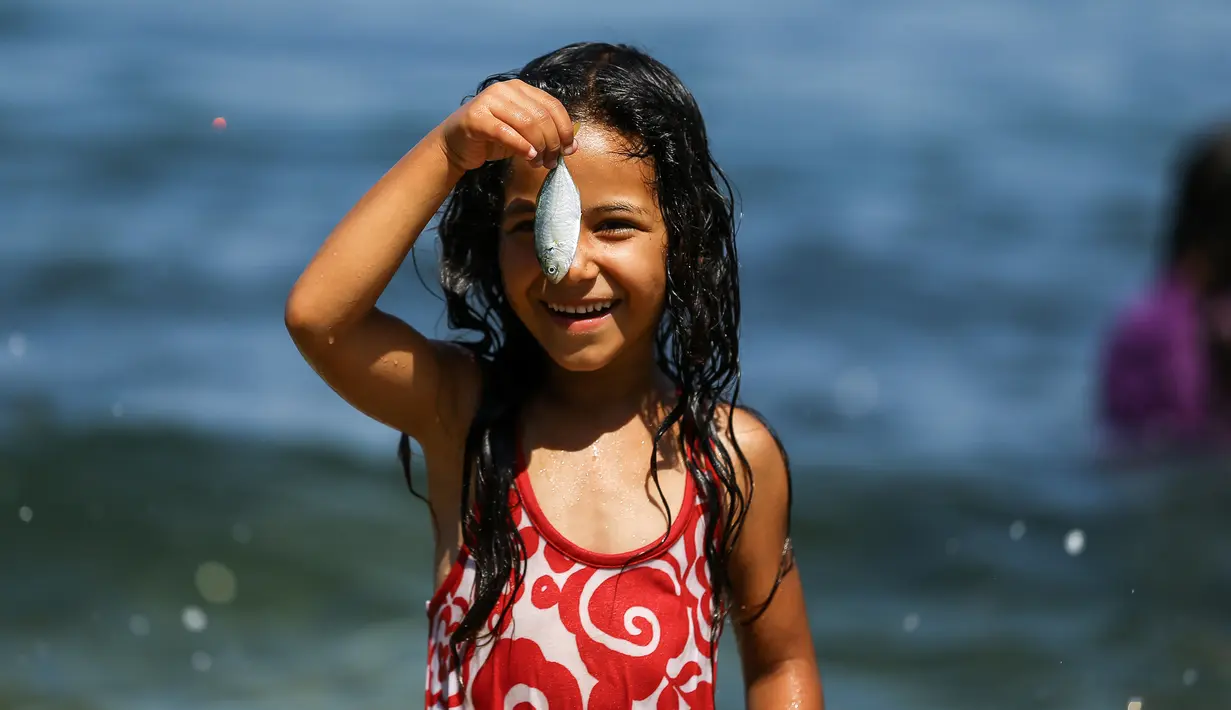 Seorang gadis Palestina tersenyum saat memegang ikan saat bermain bersama teman-temanya di laut Mediterania dekat kamp pengungsi al-Shati di Kota Gaza (3/7). (AFP Photo/Mohammed Abed)