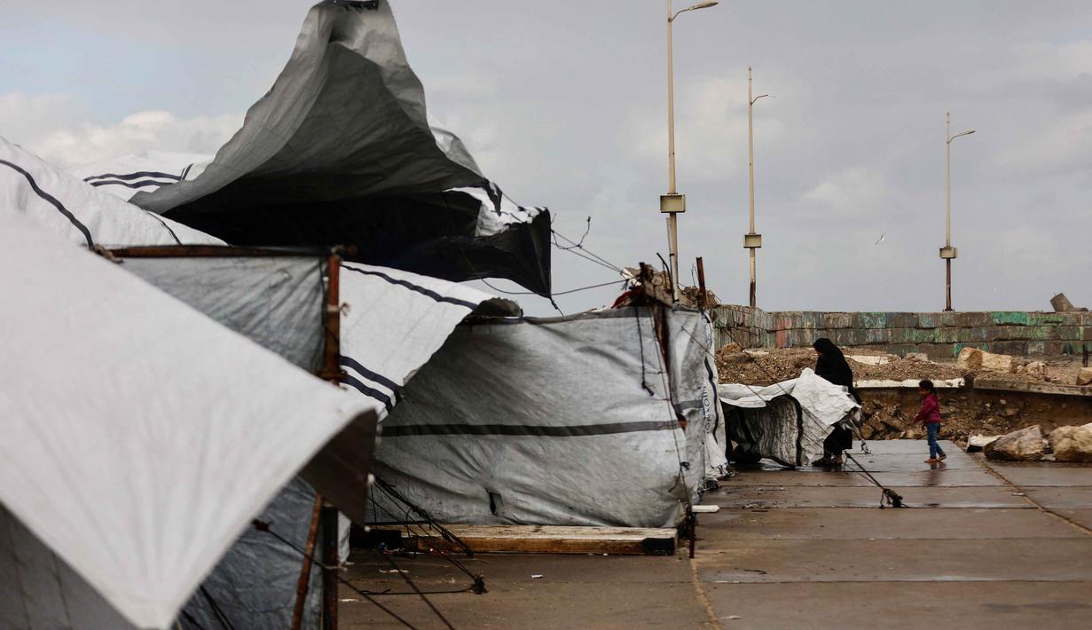 Tinggal di tenda usang dengan kondisi terpal yang compang-camping atau di tengah puing-puing bangunan. Tampak dalam foto, seorang perempuan Palestina pengungsi menyesuaikan terpal tenda keluarga saat wilayah tersebut dilanda hujan dan kondisi musim dingin yang dingin, di Kota Gaza pada 28 Desember 2025. (Omar AL-QATTAA/AFP)
