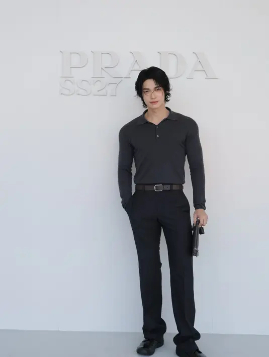 Win Metawin kenakan polo rajut abu-abu gelap lengan panjang. Dipadukan celana kain hitam, ikat pinggang cokelat, dan loafers hitam, sambil menenteng tas kulit hitam [dok. Prada]