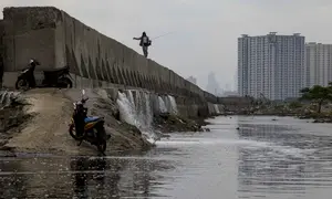 Namun, kali ini, Banjir rob di kawasan Muara Baru Jakarta diperparah dengan kebocoran dan rembesan pada tanggul laut di beberapa titik di Muara Baru, yang disebabkan oleh usia tanggul yang sudah tua dan korosi. Tampak dalam foto, seorang pria dengan pancingnya berjalan di atas tanggul laut raksasa yang dibangun untuk mencegah banjir di pesisir utara, seperti terlihat di Muara Baru, Jakarta pada Sabtu 6 Desember 2025. (ADITYA IRAWAN/AFP)