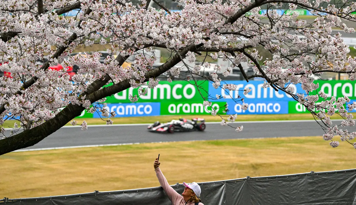 Seorang penonton mendokumentasikan foto melalui gawainya saat seorang pembalap F1 melewati pohon sakura saat melakukan latihan bebas kedua (FP2) F1 GP Jepang 2024 di Sirkuit Suzuka, Jepang, Jumat (05/04/2024). (AFP/Yuichi Yamazaki)