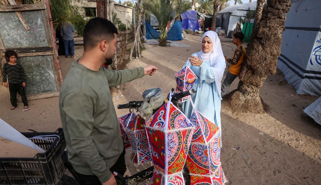 Fanous menjadi simbol suka cita, harapan, dan tradisi yang tetap bertahan di tengah situasi konflik berkepanjangan antara Israel dan Hamas. Tampak dalam foto, Rehan Hazem Sharab, seorang pengungsi Palestina berusia 32 tahun, mengantarkan lampion buatan tangannya, yang dibuat dari kotak-kotak bantuan bekas, sebagai persiapan menyambut bulan suci Ramadan yang dimulai pada 17 Februari, di luar tendanya di lingkungan Al-Mawasi, Khan Younis, di Jalur Gaza selatan pada Sabtu 7 Februari 2026. (Bashar Taleb/AFP)