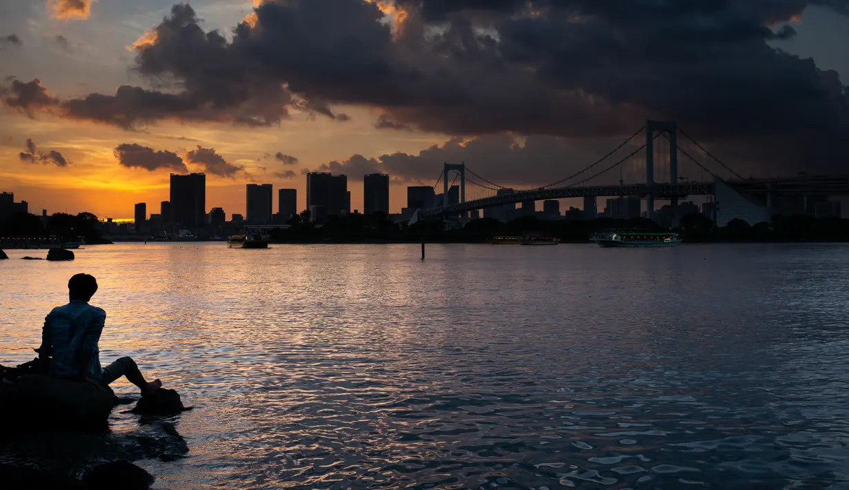 Seorang pria berada di samping The Rainbow Bridge saat matahari terbenam di Tokyo, Jepang (29/7). Odaiba merupakan salah satu tujuan wisata di Tokyo, sekaligus lokasi pusat perbelanjaan dan permukiman. (AFP Photo/Martin Bureau)