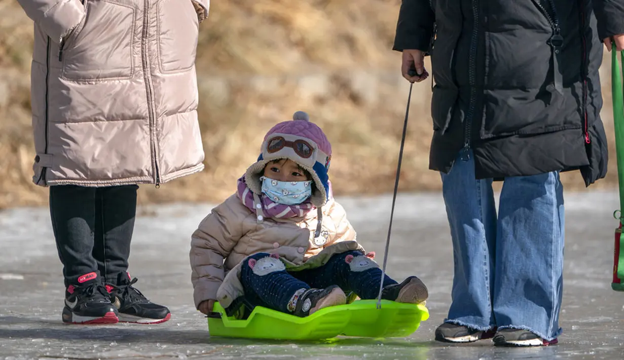 Seorang anak yang memakai masker wajah untuk melindungi dari penyebaran virus corona COVID-19 ditarik dengan kereta luncur melintasi permukaan kanal yang membeku di Beijing, China, Sabtu (30/1/2021). (AP Photo/Mark Schiefelbein)