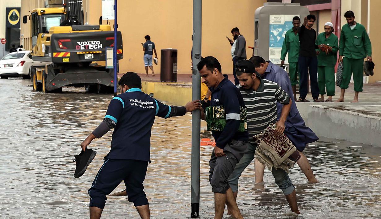 Pemerintah Dubai, UEA, mendesak warga untuk tetap berada di dalam rumah, kecuali jika benar-benar diperlukan. Tampak dalam foto, para pria mengangkat ujung celana mereka untuk menyeberangi genangan air banjir saat melintasi jalan setelah hujan lebat, di Dubai, Uni Emirat Arab (UEA) pada Jumat 19 Desember 2025. (FADEL SENNA/AFP)