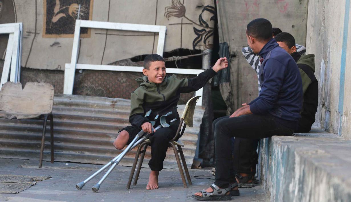 Ibu dan saudara laki-lakinya tewas dalam peristiwa pemboman tersebut. Sementara, salah satu kaki Rateb Mahmoud Qaliq terluka parah dan kemudian terpaksa harus diamputasi. Tampak dalam foto, Rateb Mahmoud Qaliq (kiri) berbincang dengan rekannya di halaman sekolah UNRWA yang telah menjadi tempat penampungan bagi pengungsi Palestina di Deir al-Balah, di Jalur Gaza tengah, pada 3 Desember 2025. (Bashar Taleb/AFP)