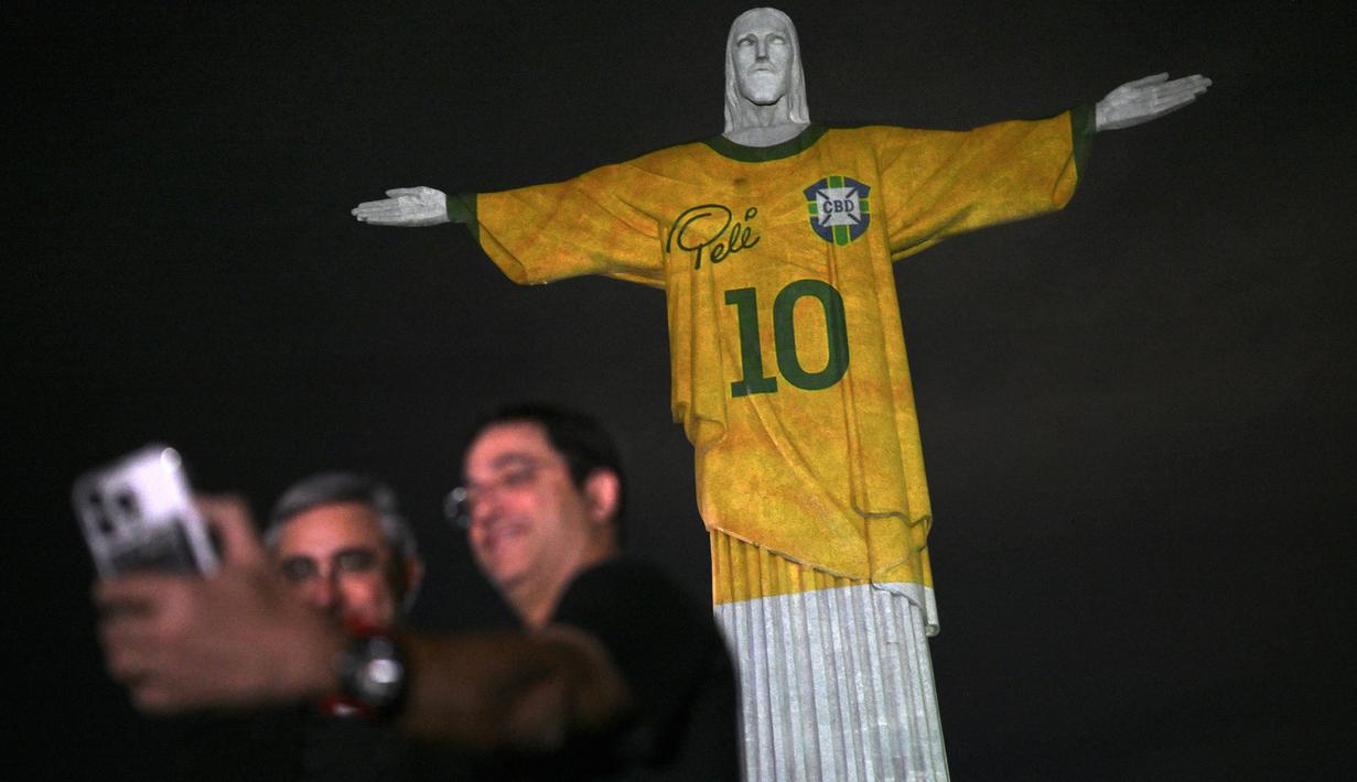Sejumlah pengunjung berfoto bersama dengan Patung Kristus Sang Penebus (Christ the Redeemer) di Rio de Janeiro, Brasil saat diterangi gambar jersey dari legenda sepak bola Brasil, Pele sebagai penghormatan atas satu tahung kematian sang pemain pada Jumat, 29 Desember 2023. (AFP/Mauro Pimentel)