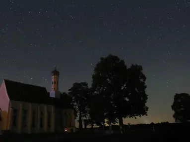 Sebuah meteor melintas di tengah langit malam selama hujan meteor tahunan Perseid di belakang gereja ziarah Sankt Coloman, barat daya Jerman, Minggu (12/8). Puncak hujan meteor terjadi pada 11-12 Agustus dan 12-13 Agustus. (KARL-JOSEF HILDENBRAND/DPA/AFP)