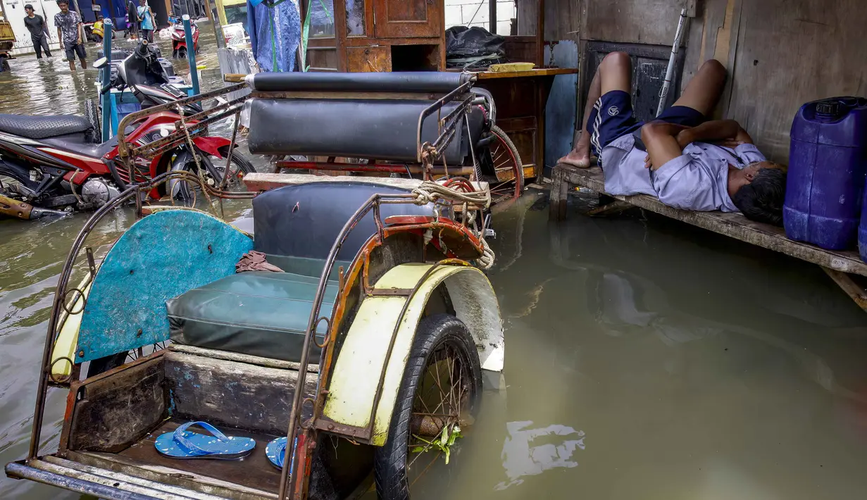 Seorang pria tertidur di samping becak, kendaraan roda tiga bertenaga manusia, sementara air pasang membanjiri jalanan di Muara Baru, Jakarta pada Sabtu 6 Desember 2025. Banjir rob akibat air pasang laut yang tinggi merendam jalanan dan pemukiman di kawasan Muara Baru, Penjaringan, Jakarta Utara sejak beberapa hari lalu, dengan puncaknya terjadi pada Jumat 5 Desember 2025. (ADITYA IRAWAN/AFP)