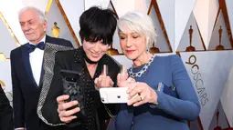 Aktris senior Helen Mirren melakukan wefie dengan sahabat sekaligus penyanyi Diane Warren di karpet merah Piala Oscar 2018, Los Angeles, Minggu (4/3). Acungan jari tengahnya tersebut menjadi perbincangan di internet. (Christopher Polk/GETTY IMAGES/AFP)