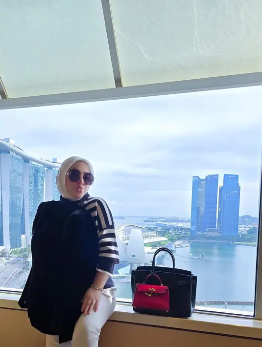 <p>Di foto lainnya, Syahrini juga masih menutup bagian perutnya dengan outer. Wanita berhijab itu berpose di kaca dengan pemandangan Marina Bay Sands. (foto: Instagram/ princessyahrini)</p>