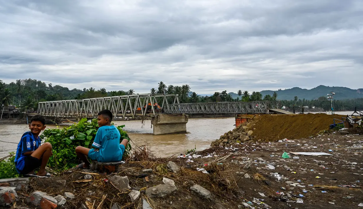 Pihak terkait akan terus mengerahkan kemampuan untuk melakukan upaya percepatan pemulihan infrastruktur dan konektivitas wilayah terdampak bencana. Tampak dalam foto, anak-anak duduk di dekat jembatan yang rusak akibat banjir di Juli, provinsi Aceh, pada Minggu 14 Desember 2025. (CHAIDEER MAHYUDDIN/AFP)