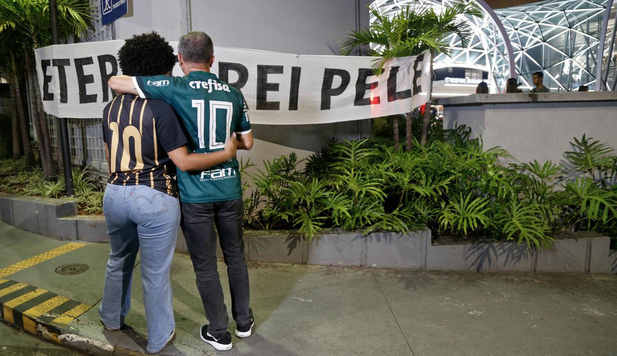 Dua orang berdiri di depan spanduk bertuliskan "Raja Abadi Pele" di luar Rumah Sakit Israel Albert Einstein, tempat legenda sepak bola Brasil itu meninggal setelah lama berjuang melawan kanker di Sao Paulo, Brasil, pada 29 Desember 2022. (AFP/Miguel Schincariol)