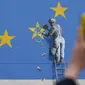 Warga mengambil gambar mural seorang pria yang tengah menghancurkan salah satu dari 12 bintang kuning bendera Uni Eropa di dinding kawasan Dover, Inggris, Senin (8/5). Mural karya seniman jalanan Banksy itu berjudul 'Brexit'. (DANIEL LEAL-OLIVAS/AFP)