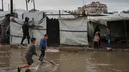 Air terlihat menggenangi jalanan dan memasuki tenda. Tampak dalam foto, anak-anak pengungsi Palestina berjalan melewati kamp tenda yang basah kuyup akibat hujan lebat di Deir al-Balah, Jalur Gaza tengah, Kamis, 26 Maret 2026. (AP Photo/Abdel Kareem Hana)