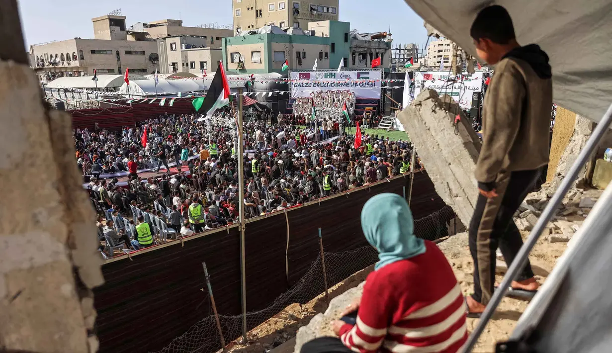 Kegiatan ini digelar di tengah bayang-bayang blokade dan kondisi kemanusiaan yang masih berlangsung di Gaza. Tampak dalam foto, seorang anak laki-laki dan perempuan mengamati dari sebuah bangunan yang rusak parah saat orang-orang berkumpul selama perayaan pernikahan massal untuk para pengantin Palestina yang diselenggarakan oleh Yayasan Bantuan Kemanusiaan Turki (IHH) di Kota Gaza pada 11 Mei 2026. (Omar AL-QATTAA/AFP)