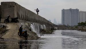 Namun, kali ini, Banjir rob di kawasan Muara Baru Jakarta diperparah dengan kebocoran dan rembesan pada tanggul laut di beberapa titik di Muara Baru, yang disebabkan oleh usia tanggul yang sudah tua dan korosi. Tampak dalam foto, seorang pria dengan pancingnya berjalan di atas tanggul laut raksasa yang dibangun untuk mencegah banjir di pesisir utara, seperti terlihat di Muara Baru, Jakarta pada Sabtu 6 Desember 2025. (ADITYA IRAWAN/AFP)