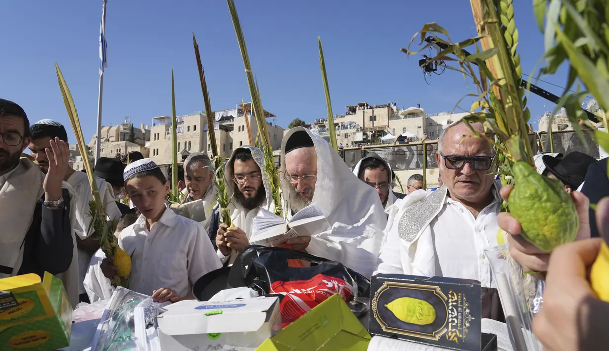 Para penyembah berdoa dengan empat spesies Sukkot — cabang palem, myrtle, willow dan etrog, buah jeruk, ketika pria Yahudi ultra-Ortodoks dari kasta Cohanim Priestly berdoa dengan ditutupi selendang doa saat mereka berpartisipasi dalam pemberkatan selama hari raya Yahudi Sukkot di Tembok Barat, situs paling suci di mana orang Yahudi bisa berdoa, di Kota Tua Yerusalem Rabu, 12 Oktober 2022. (AP Photo/Tsafir Abayov)