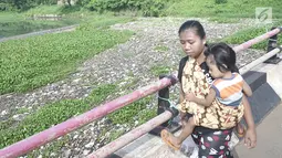 Seorang ibu menggendong anaknya berjalan melintasi Setu Pengarengan yang dipenuhi sampah dan eceng gondok di Depok, Jawa Barat, Selasa (7/5/2019). Kurangnya perawatan menyebabkan Setu Pengarengan kian memprihatinkan. (Liputan6.com/Immanuel Antonius)