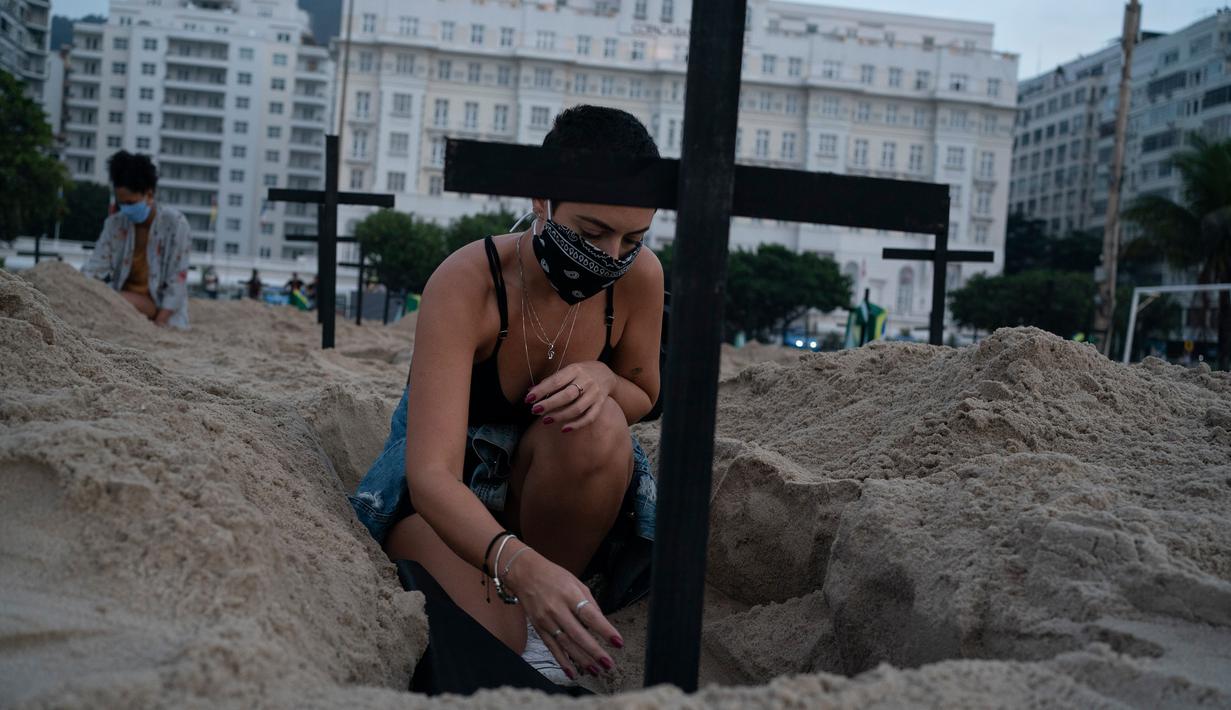 Seorang aktivis meletakkan salib di kuburan yang digali sebagai bentuk protes di pantai Copacabana, Rio de Janeiro, Kamis (11/6/2020). Kuburan-kuburan yang dihiasi bendera Brasil kecil itu untuk mengenang puluhan ribu pasien corona yang meninggal di negara tersebut. (AP Photo/Leo Correa)