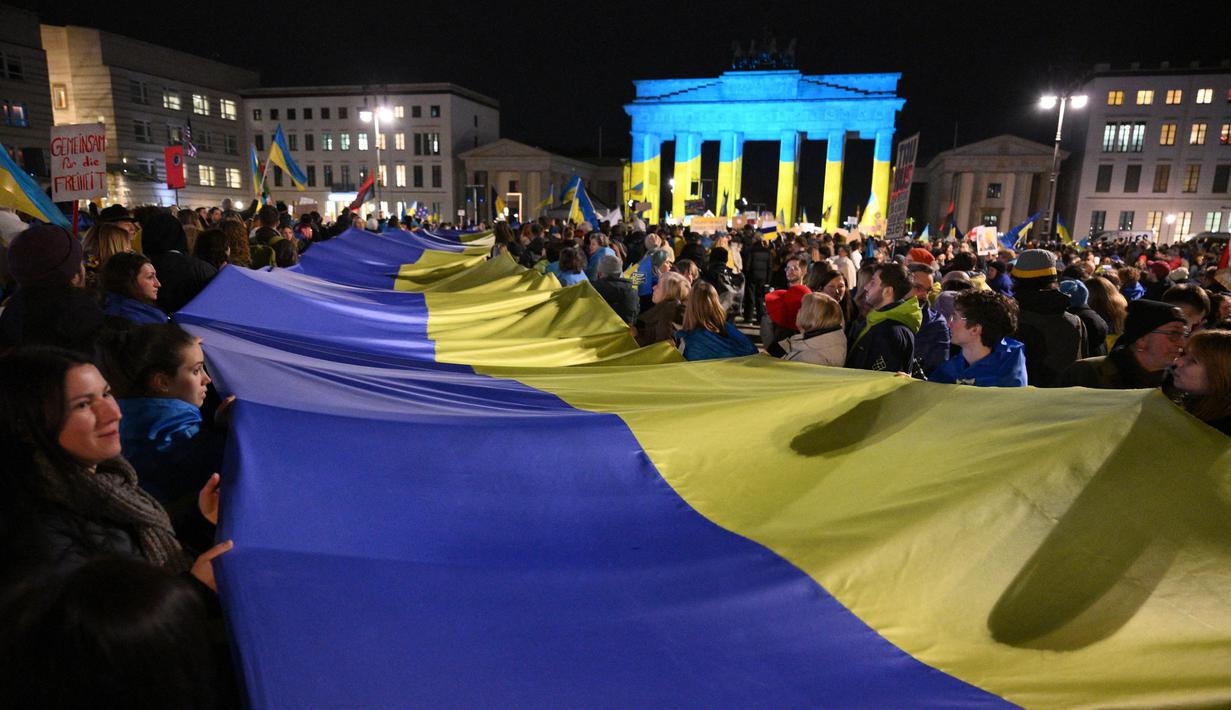 Orang-orang mengambil bagian dalam unjuk rasa pro-Ukraina di depan Gerbang Brandenburg, yang diterangi dengan warna-warna bendera Ukraina, menandai tiga tahun invasi Rusia ke Ukraina di Berlin pada 24 Februari 2025. (RALF HIRSCHBERGER/AFP)