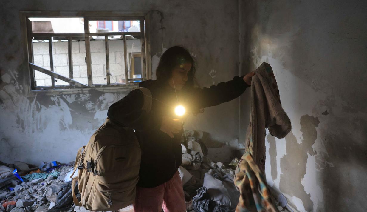 Sementara, jumlah korban luka meningkat drastis hingga melebihi 171.324 orang. Tampak dalam foto, seorang wanita Palestina menggunakan senter di ponselnya untuk mengambil pakaian dari bawah reruntuhan di sebuah ruangan, setelah serangan militer Israel terhadap rumah keluarga al-Houli, di mana empat orang dilaporkan tewas, di sebelah barat Deir al-Balah, di Jalur Gaza tengah pada Jumat 16 Januari 2026. (BASHAR TALEB/AFP)