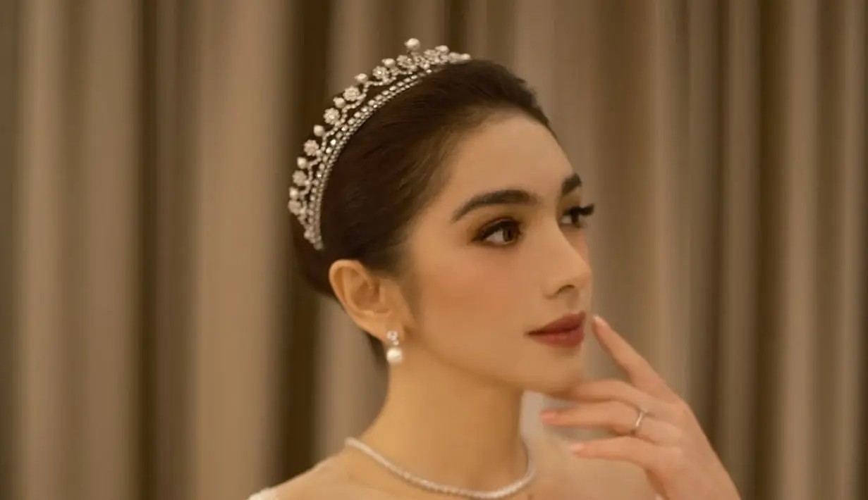 Gaun pengantin yang dikenakan Angel Karamoy memiliki potongan square neckline yang elegan dengan lengan panjang berbahan lace transparan. Siluet ball gown yang mengembang dengan ekor panjang menambah kesan mewah bak putri kerajaan. Aksesori tiara kristal dan anting mutiara semakin mempercantik tampilannya. (Liputan6.com/IG/@realangelkaramoy)