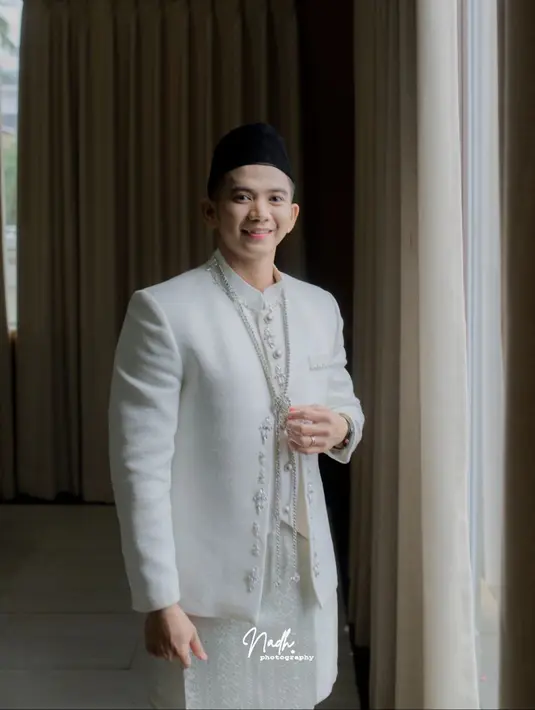 Rizki DA tampak mengenakan atasan dan bawahan serba putih yang dilapisi sarung pendek. Ia melengkapi penampilannya dengan peci dan sepatu pantofel hitam. [@da2_rizki123/@nadh.photography]