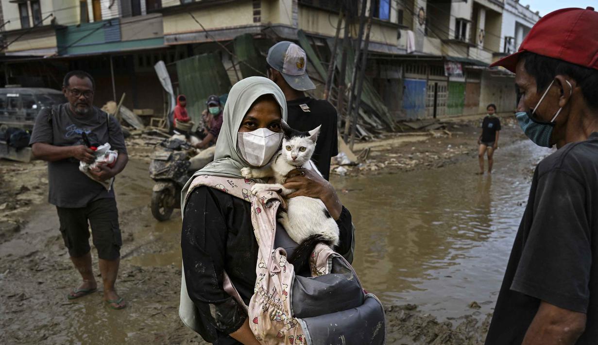 Gelombang penyakit pertama yang mengancam adalah diare, infeksi kulit, gangguan pencernaan, hingga penyakit pernapasan. Tampak dalam foto, seorang perempuan menggendong kucingnya di jalanan yang masih berlumpur setelah banjir bandang melanda daerah tersebut di Aceh Tamiang, Provinsi Aceh, pada Sabtu 6 Desember 2025. (YT HARIONO/AFP)