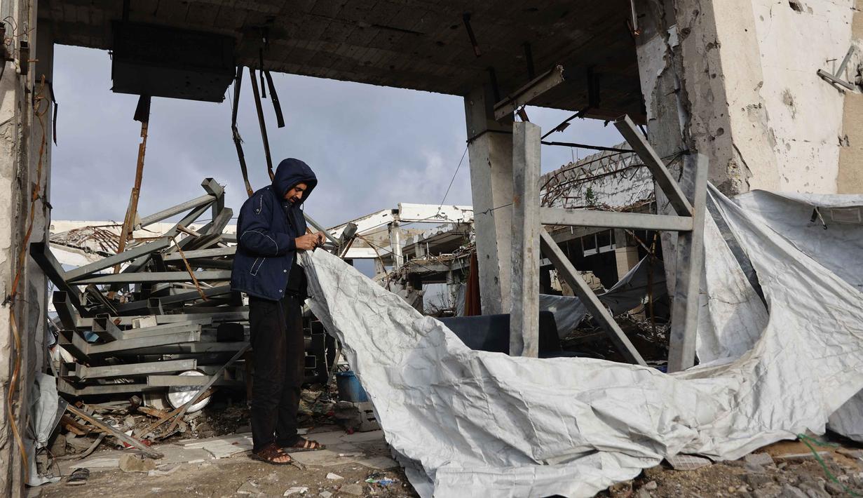 Warga Palestina di Gaza terpaksa bertahan di tenda-tenda yang rusak atau rumah-rumah yang tidak layak huni karena proses pembangunan kembali terhambat. Tampak dalam foto, seorang pria Palestina memperbaiki tenda perlindungan di sebuah bangunan yang rusak akibat perang, dimana sebagian bangunan tersebut runtuh pada hari musim dingin yang berangin di Kota Gaza pada Selasa 13 Januari 2026. (Omar AL-QATTAA/AFP)