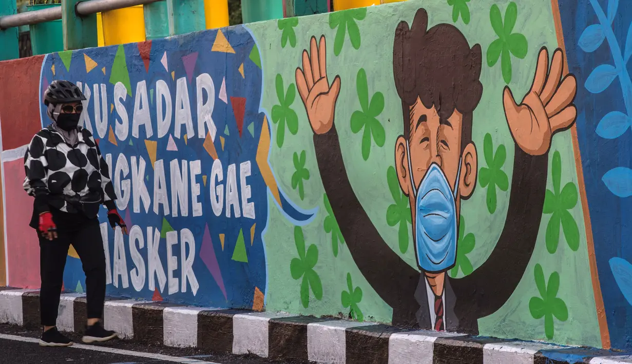 Seorang perempuan melewati mural yang mengajak orang untuk memakai masker di tengah pandemi Covid-19 di Surabaya, Minggu (25/10/2020). Mural di sepanjang dinding itu sebagai sarana imbauan kepada masyarakat untuk menerapkan protokol kesehatan pencegahan penularan COVID-19. (Juni Kriswanto/AFP)