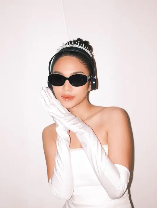 Ia tampil totalitas mengenakan mini dress putih, lengkap dengan sarung tangan, mahkota, headphone, dan kacamata hitam sebagai aksesori.  [@azizahsalsha_]
