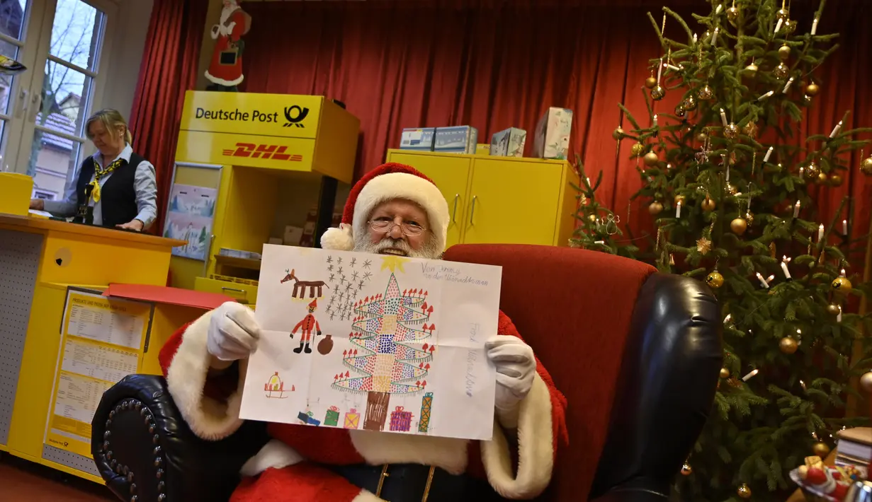 Pria berkostum Sinterklas menunjukkan hadiah lukisan dari seorang anak di Kantor Pos Natal di Himmelpfort, Jerman, 19 Desember 2018. Anak-anak di seluruh dunia mulai mengirim surat berisi permohonan kepada sinterklas di kantor pos itu (Tobias SCHWARZ/AFP)