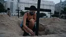 Seorang aktivis meletakkan salib di kuburan yang digali sebagai bentuk protes di pantai Copacabana, Rio de Janeiro, Kamis (11/6/2020). Kuburan-kuburan yang dihiasi bendera Brasil kecil itu untuk mengenang puluhan ribu pasien corona yang meninggal di negara tersebut. (AP Photo/Leo Correa)