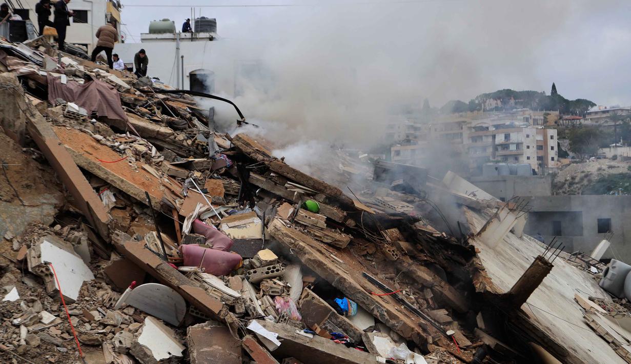 Israel berdalih, serangan tersebut bertujuan untuk menghancurkan fasilitas militer dan gudang senjata Hizbullah guna mencegah ancaman di sepanjang perbatasan. Tampak dalam foto, asap mengepul saat orang-orang memeriksa bangunan yang hancur akibat serangan Israel di desa Qennarit, Lebanon selatan, Kamis 22 Januari 2026. (AP Photo/Mohammed Zaatari)