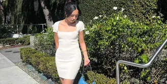 Dalam foto ini Beby Tsabina tampil kenakan dress putih yang kasual. Berpose membelakangi laut, Beby Tsabina menyempurnakan gayanya dengan menenteng tas Prada, menata rambutnya dengan gaya high-bun, dan mengenakan sneakers putih. Foto: Instagram.
