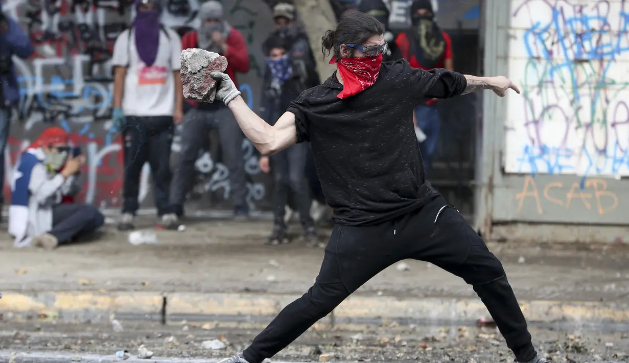 Seorang demonstran anti-pemerintah melemparkan batu ke arah polisi di Santiago, Chile (4/10/2019). Pengunjuk rasa menuntut diakhirinya ketidaksetaraan ekonomi bahkan ketika pemerintah mengumumkan bahwa demonstrasi selama berminggu-minggu merugikan pertumbuhan ekonomi negara. (AP Photo/Esteban Felix)