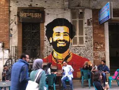 Foto file pada 4 April 2018 menunjukkan orang-orang duduk di sebuah kafe di pusat kota Kairo dengan latar belakang lukisan mural yang menggambarkan striker Liverpool, Mohamed Salah. Federasi Sepak Bola Mesir melaporkan, Jumat (13/11/2020), Mohamed Salah dinyatakan positif Covid-19. (Amir MAKAR/AFP)