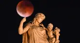 Foto ini menampilkan bulan purnama yang juga dikenal sebagai “Blood Moon” selama gerhana bulan, dengan patung dewi Yunani kuno Irene yang memegang seorang anak di latar depan, di pusat Athena, Yunani pada 7 September 2025. (Angelos TZORTZINIS/AFP)