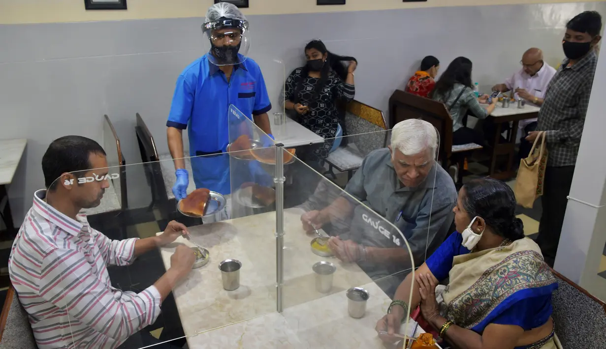 Pramusaji mengenakan pelindung wajah dan masker saat menyajikan makanan kepada pelanggan di restoran yang dibuka kembali usai pelonggaran lockdown di Bangalore, India, Senin (8/6/2020). Untuk keamanan pelanggan, restoran-restoran menyiapkan panel transparan di meja makan. (Manjunath Kiran/AFP)